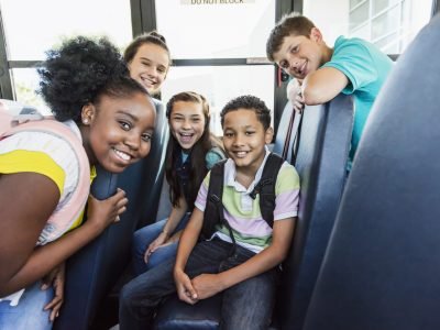 A multi-ethnic group of five junior high or middle school students, 11 to 13 years old, riding a school bus. They are smiling at the camera.